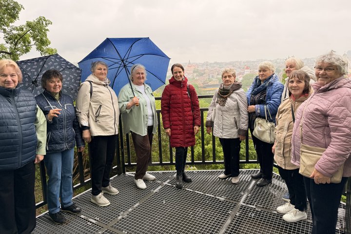 Trečiojo amžiaus universiteto studentai lietingą dieną Užupyje klausosi pasakojimo Altanos bokštelyje, kopia laiptais į viršų ir fotografuojasi tarp žydinčių alyvų.