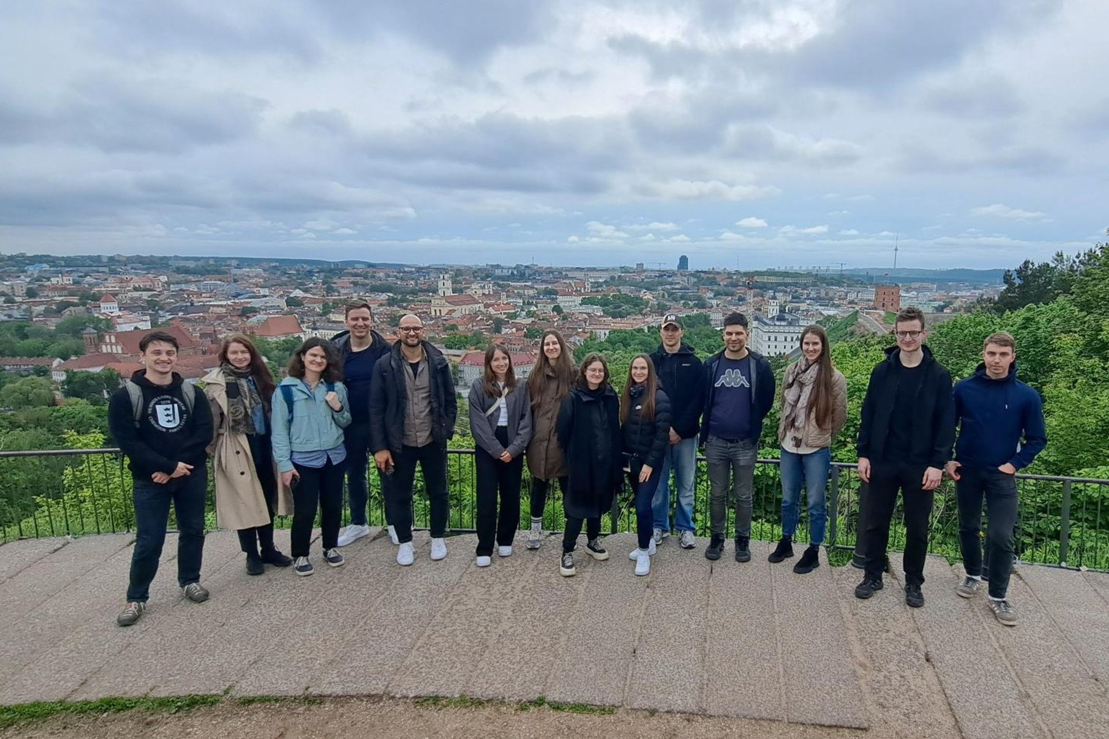 Ekskursijos dalyviai fotografuojasi apžvalgos aikštelėje Vilniaus pilių teritorijoje. Fone atsiveria Vilniaus senamiesčio panorama, apsupta žaliuojančių medžių ir debesuoto dangaus.