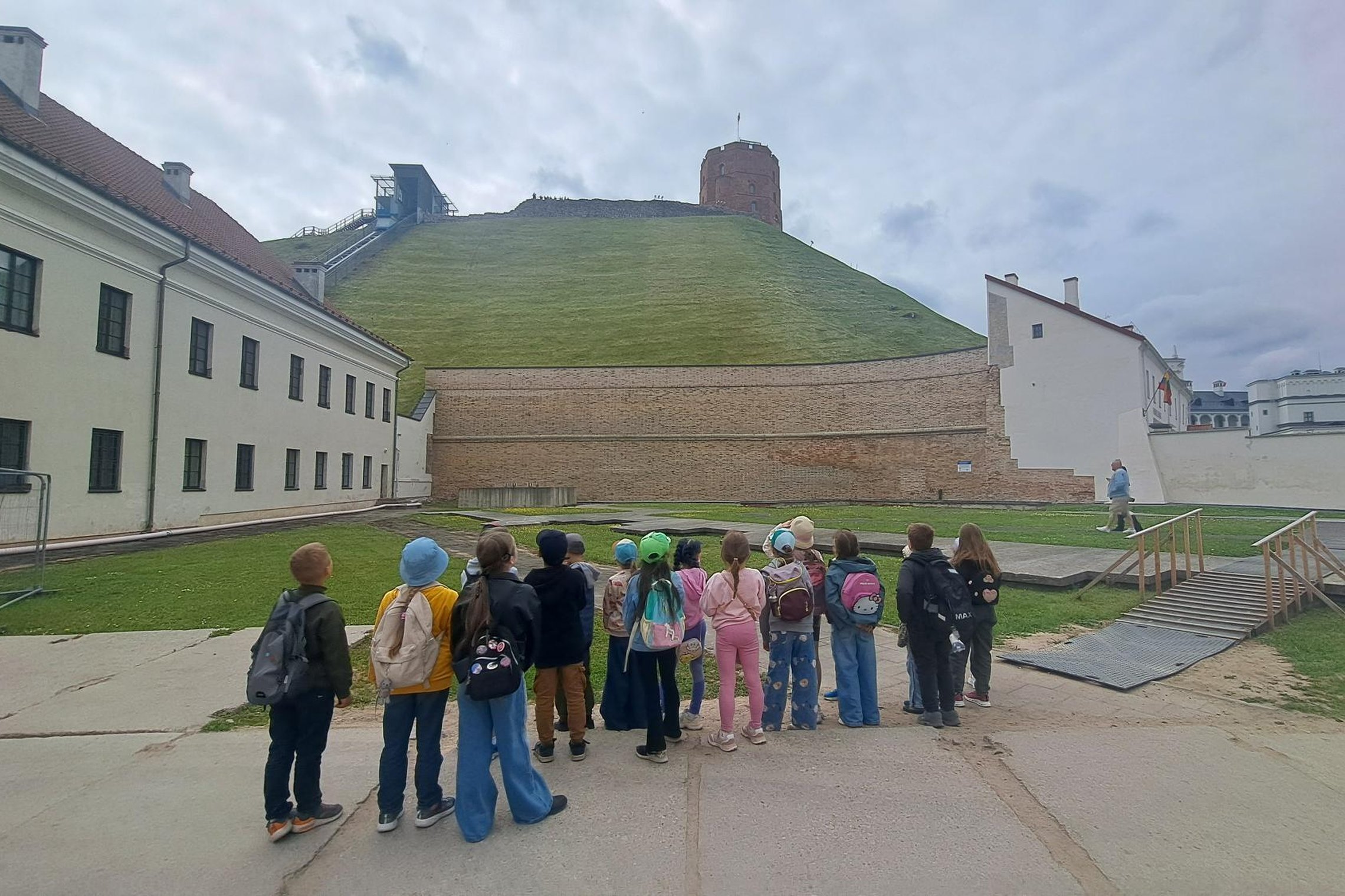 Pradinukų grupė dalyvauja edukacijoje Vilniaus pilių direkcijos teritorijoje. Vaikai stovi Žemutinės pilies teritorijoje ir klausosi edukatoriaus pasakojimo, o fone matyti Gedimino kalnas su Aukštutinės pilies Gedimino bokštu. Virš kalno kaupiasi debesuot