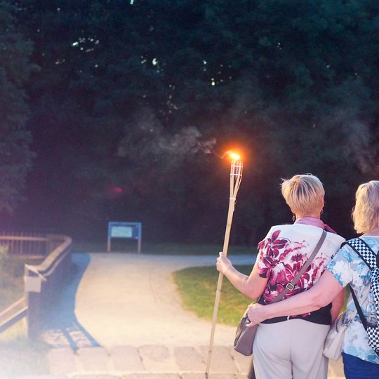 Du ekskursijos dalyviai, nešini deglais, sutemose leidžiasi Kalnų parko taku ir keliauja į vakarinę ekskursiją po Vilniaus pilių legendas. Deglų šviesa apšviečia taką ir sukuria paslaptingą atmosferą istoriniame Vilniaus pilių kraštovaizdyje.