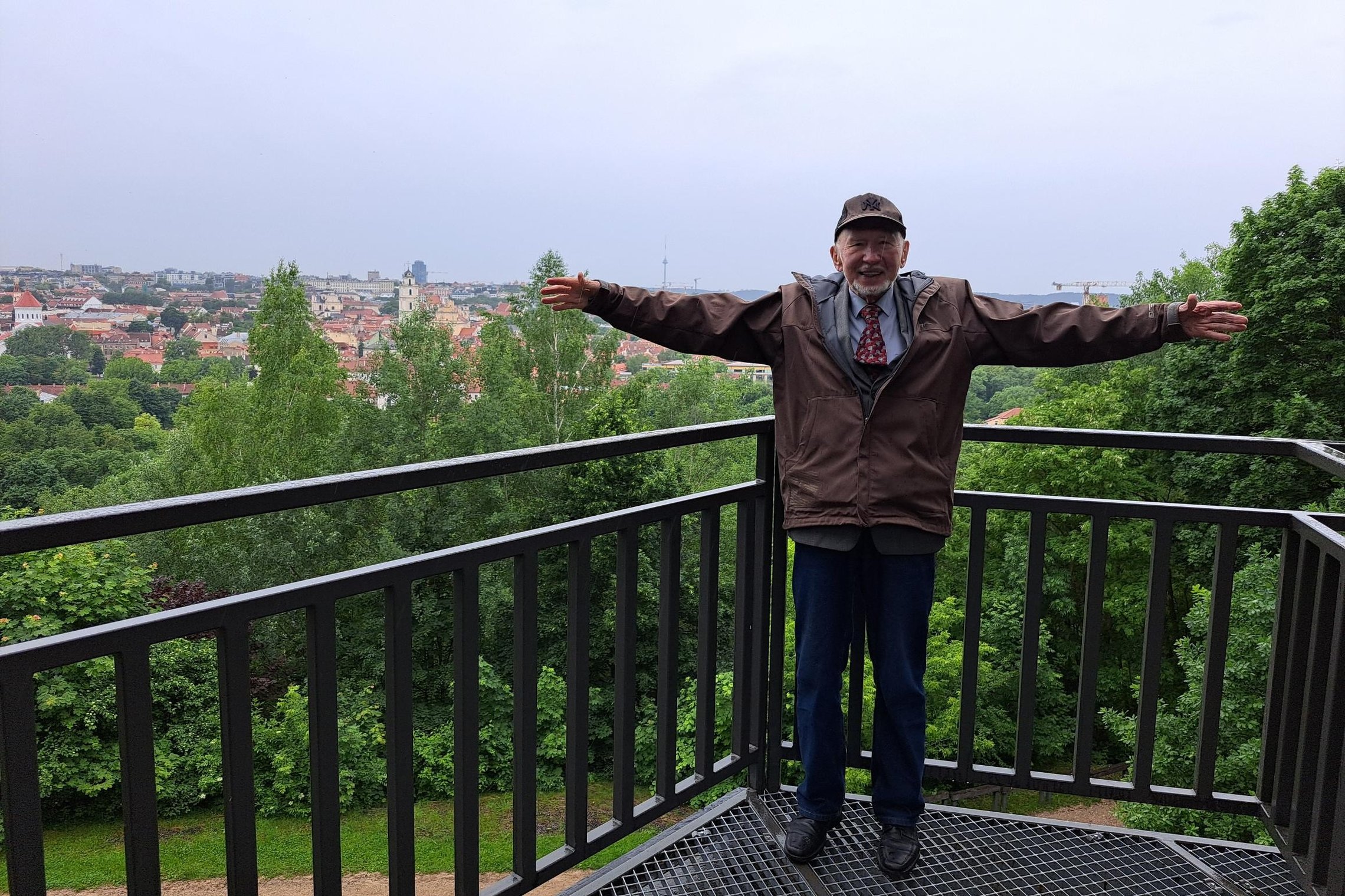Nuotraukoje Prof. Libertas Klimka stovi ant Altanos kalno bokštelio apžvalgos aikštelėje. Už jo atsiveria Vilniaus senamiesčio panorama ir žaliuojančios istorinių Vilniaus pilių kalvos.