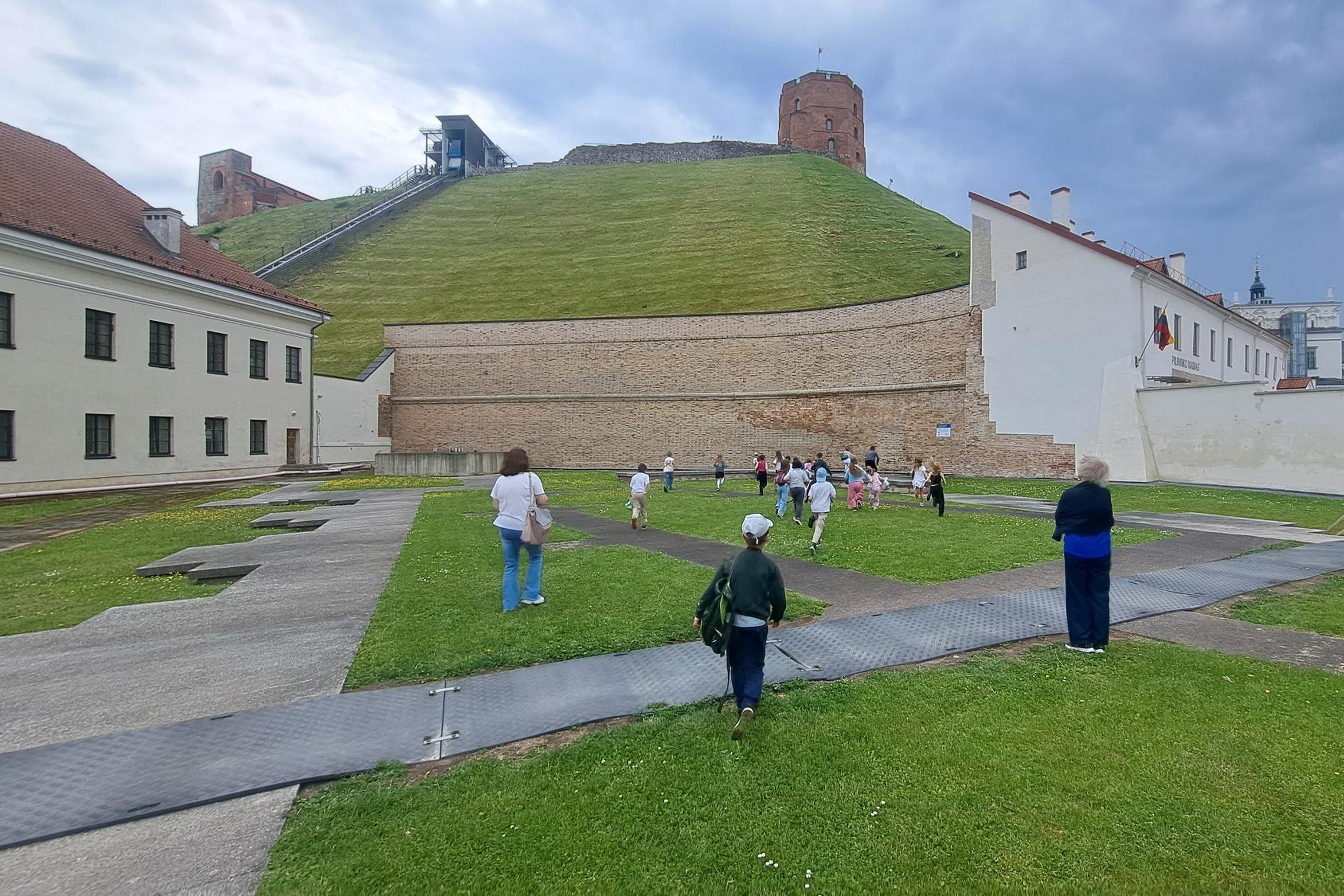 Vilniaus pilių teritorijoje vykstančios edukacijos metu vaikai su lydinčiais suaugusiaisiais eina per Žemutinės pilies kiemą Gedimino kalno link. Virš kalno stūkso Gedimino pilies bokštas, o šalia matyti funikulierius. Danguje kaupiasi tamsūs lietaus debe