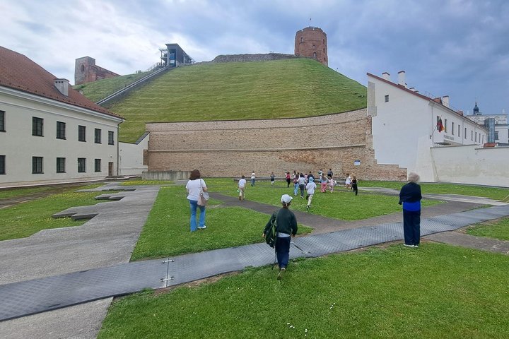 Vilniaus pilių teritorijoje vykstančios edukacijos metu vaikai su lydinčiais suaugusiaisiais eina per Žemutinės pilies kiemą Gedimino kalno link. Virš kalno stūkso Gedimino pilies bokštas, o šalia matyti funikulierius. Danguje kaupiasi tamsūs lietaus debe