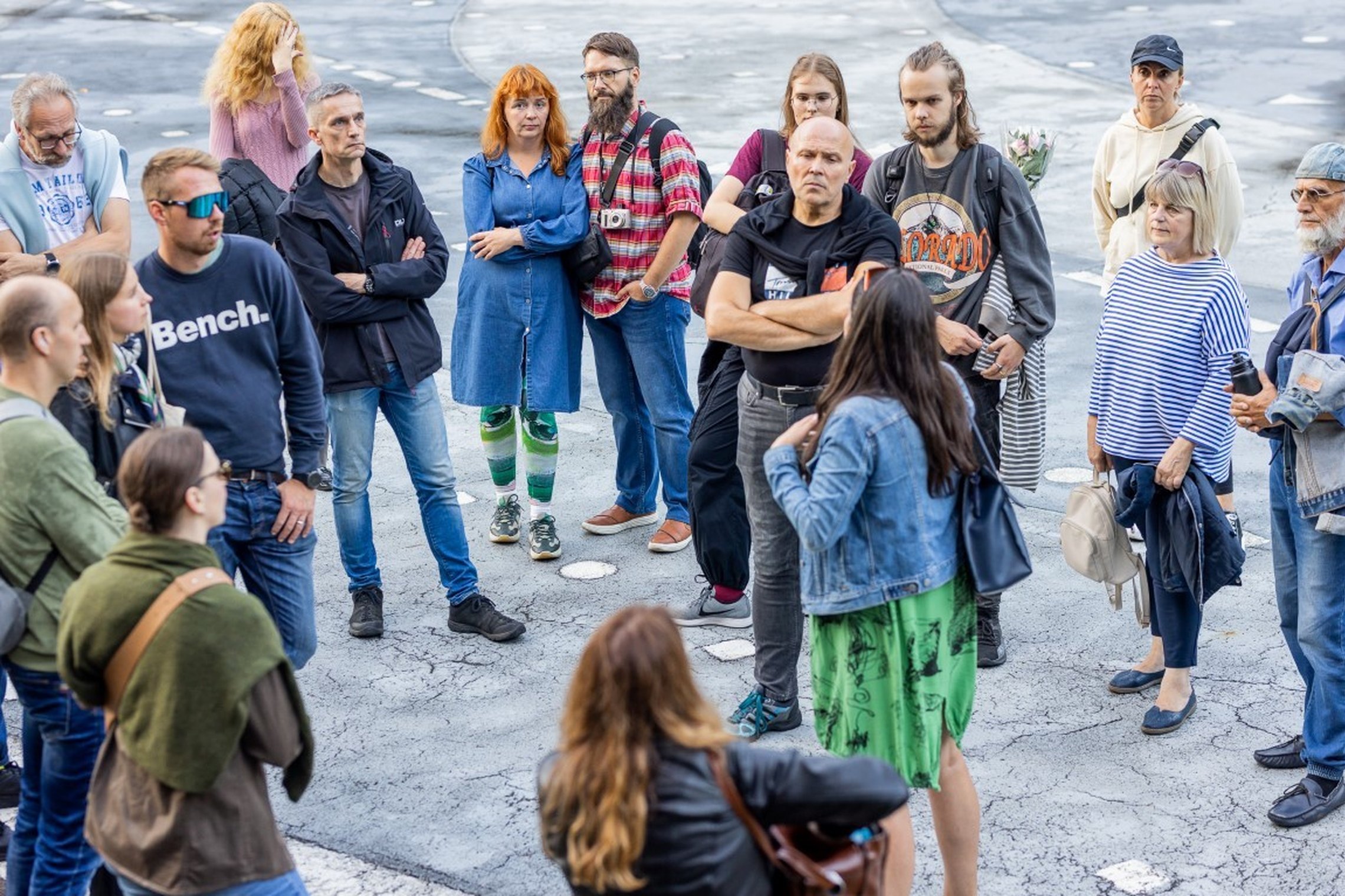 Žmonių grupė klausosi gidės pasakojimo lauke, Vilniaus pilių teritorijoje. Atidžiai susibūrę ekskursijos dalyviai stovi ratu ant pilko grindinio.