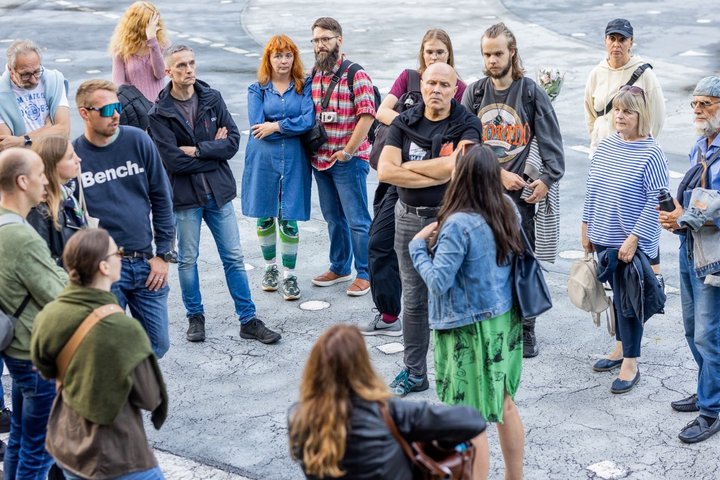 Žmonių grupė klausosi gidės pasakojimo lauke, Vilniaus pilių teritorijoje. Atidžiai susibūrę ekskursijos dalyviai stovi ratu ant pilko grindinio.