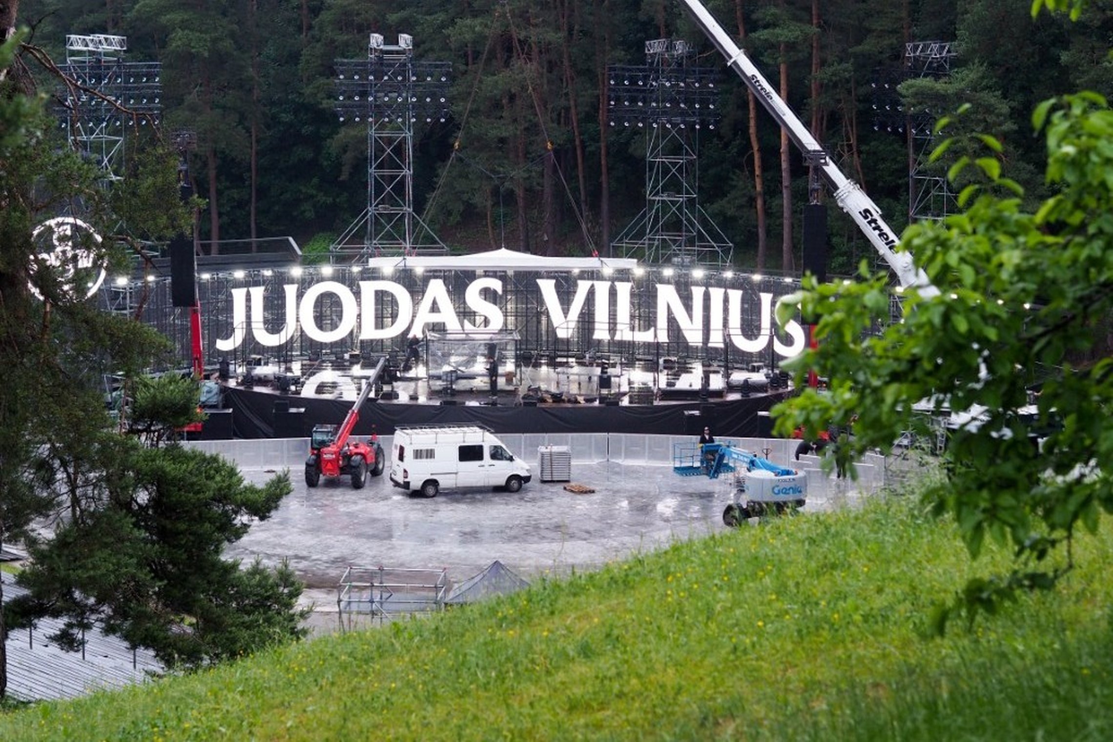 Vaizdas į Kalnų parke statomą renginio sceną, kurioje dominuoja didelis užrašas „Juodas Vilnius“. Prie scenos dirba keltuvai ir transporto priemonės, o renginio infrastruktūra įrengta miško apsuptyje.