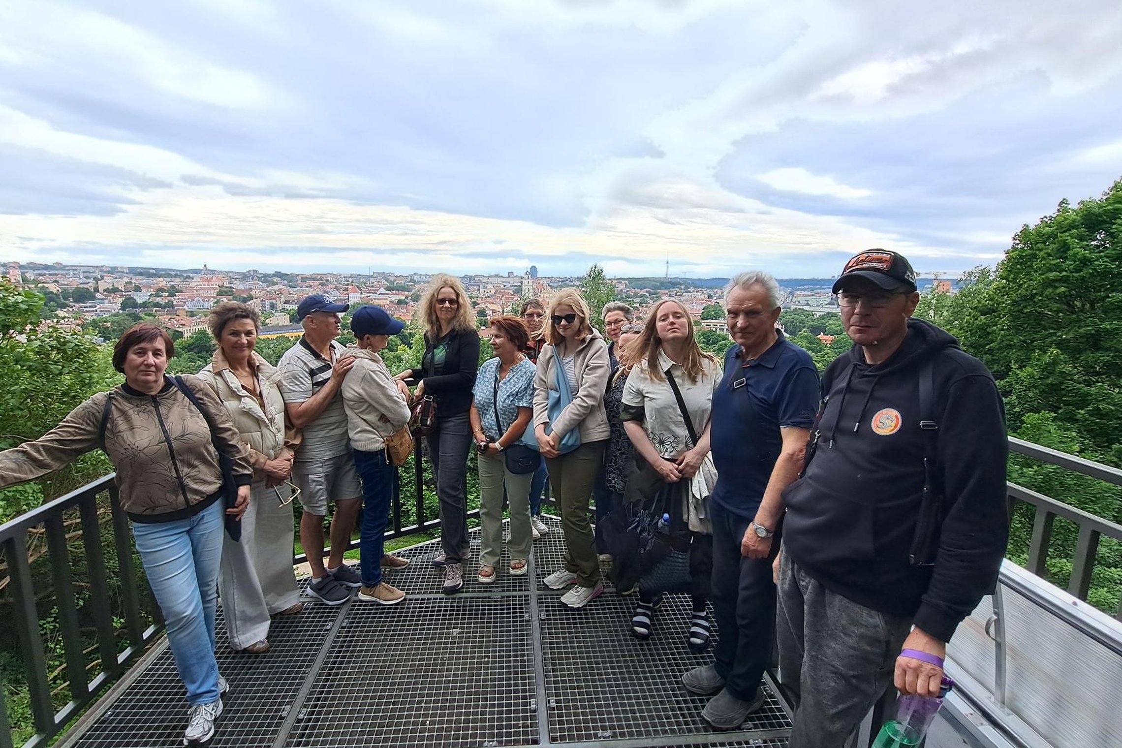 Ekskursijos dalyviai stovi ant Altanos bokštelio apžvalgos aikštelės Vilniaus pilių teritorijoje. Už jų atsiveria plati Vilniaus senamiesčio panorama, o aikštelę supa vešlūs medžiai ir žaliuojantys šlaitai.