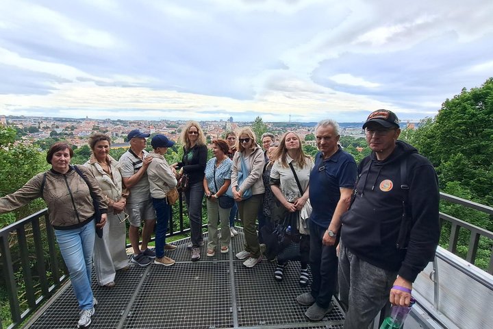 Ekskursijos dalyviai stovi ant Altanos bokštelio apžvalgos aikštelės Vilniaus pilių teritorijoje. Už jų atsiveria plati Vilniaus senamiesčio panorama, o aikštelę supa vešlūs medžiai ir žaliuojantys šlaitai.