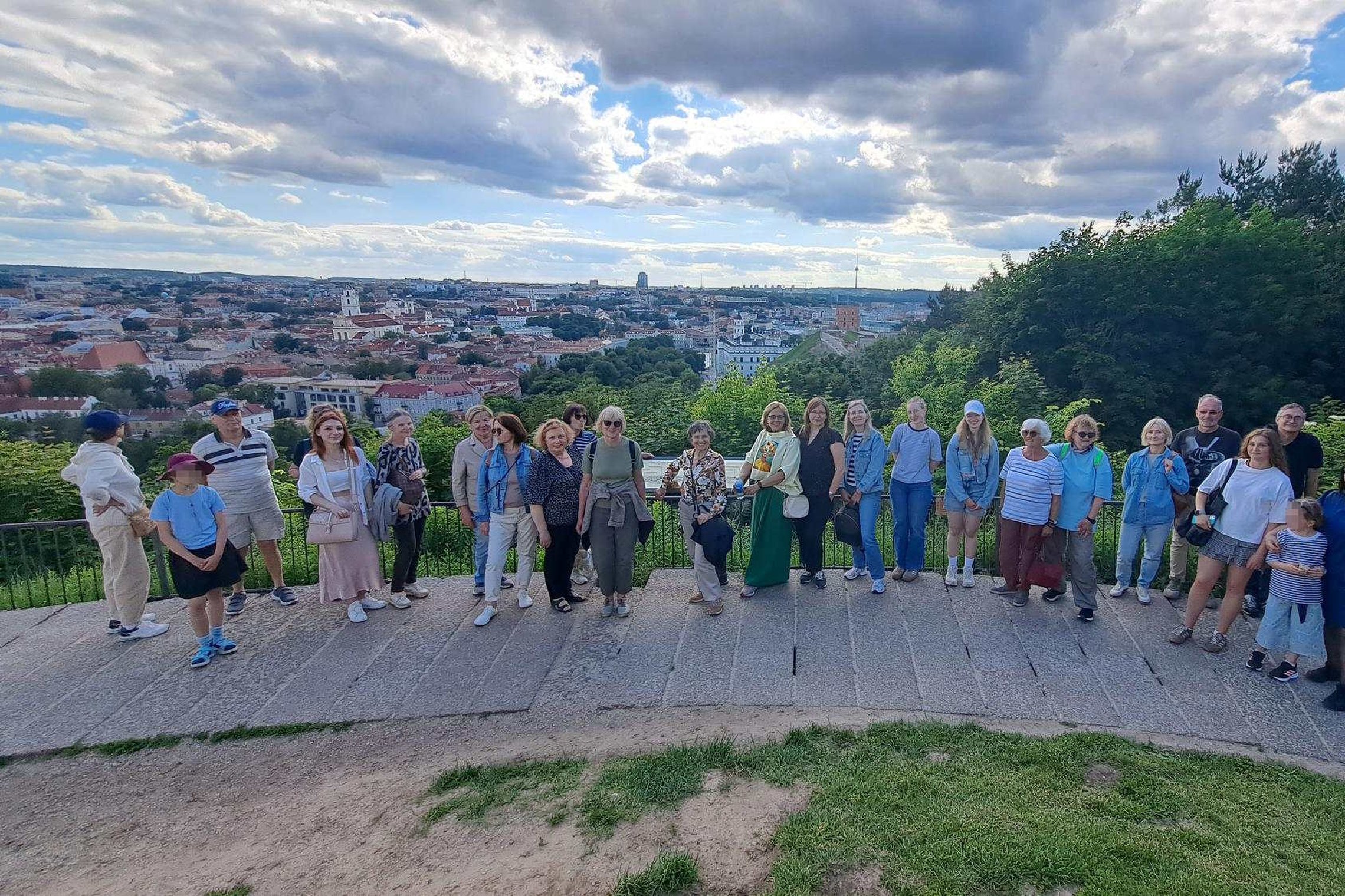Ekskursijos „Sostų karai: Gediminačių žūtbūtinis mūšis“ dalyviai stovi ant apžvalgos aikštelės Gedimino kalne. Fone atsiveria plati Vilniaus senamiesčio panorama su bažnyčių bokštais ir miesto pastatais, virš kurių driekiasi debesuotas vasaros dangus. Gru