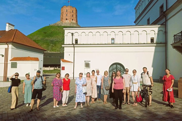 Ekskursijos „Tvanas Vilniaus pilyse“ dalyviai stovi Vilniaus pilių teritorijoje, Gedimino pilies bokšto fone, saulėtą karštą dieną.