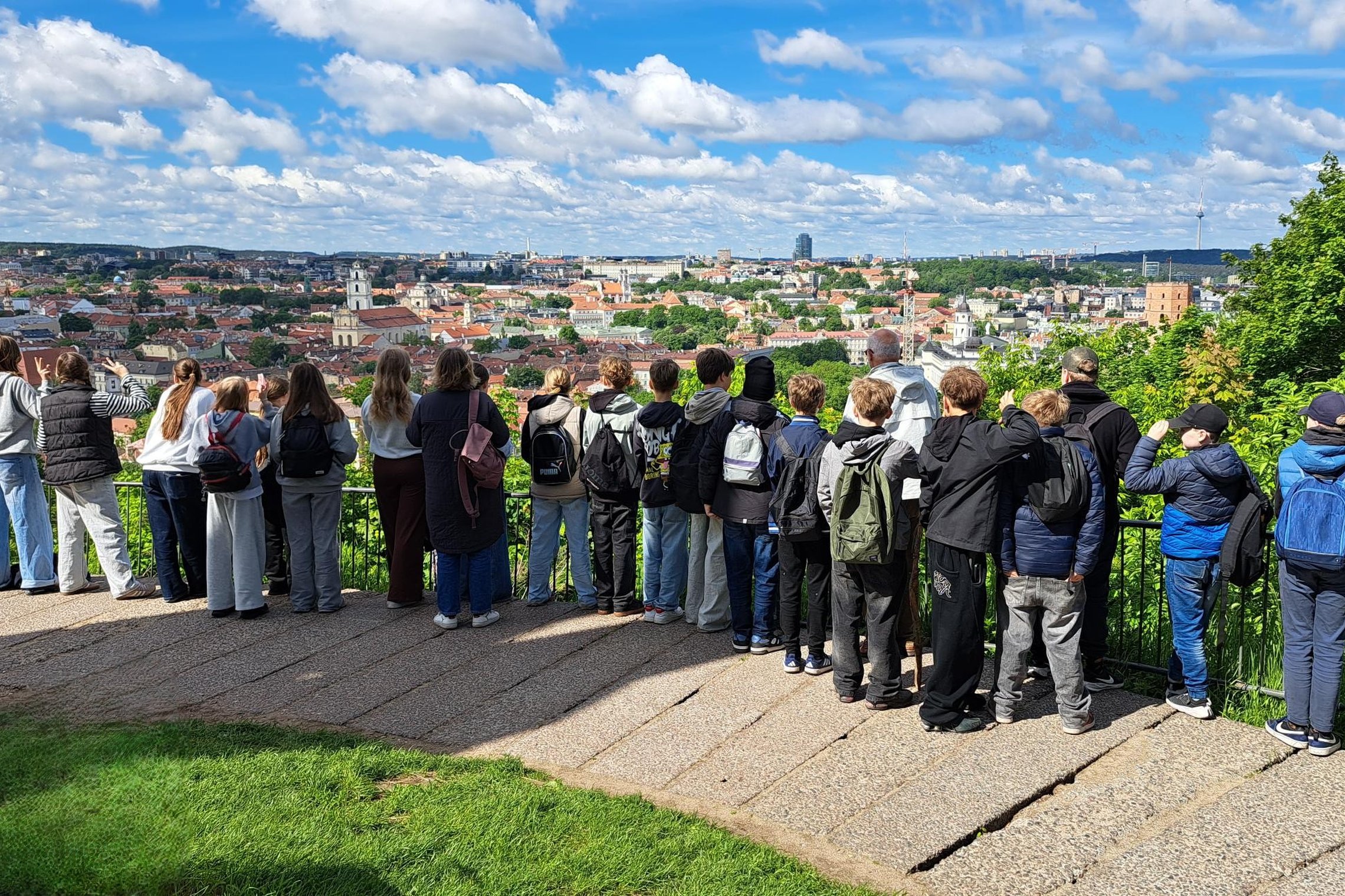 Vilniaus Martino Mažvydo progimnazijos ir Šiuolaikinės mokyklos centro penktokai stovi Vilniaus pilių teritorijos apžvalgos aikštelėje ir žvelgia į Vilniaus senamiesčio panoramą. Fone matyti miesto bokštai, raudoni stogai ir žaliuojančios kalvos.