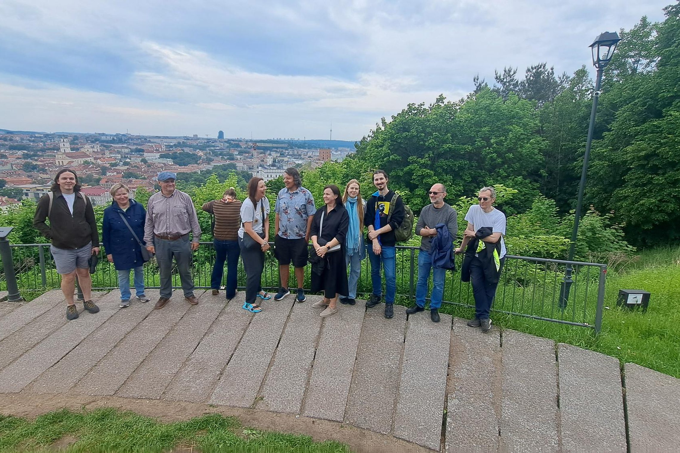 Ekskursijos dalyviai stovi ant apžvalgos aikštelės prie Vilniaus pilių teritorijos. Už jų atsiveria plati Vilniaus senamiesčio panorama su bažnyčių bokštais ir miesto stogais, o dešinėje pusėje matyti vešli žaluma ir medžiais apaugęs šlaitas. Debesuotas d