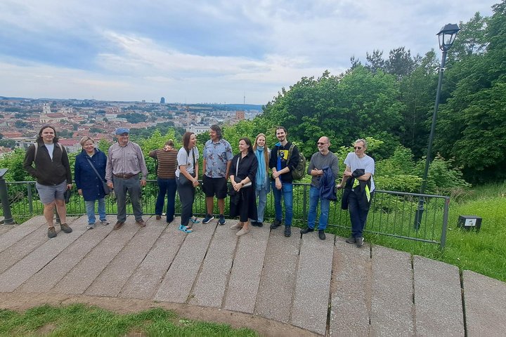 Ekskursijos dalyviai stovi ant apžvalgos aikštelės prie Vilniaus pilių teritorijos. Už jų atsiveria plati Vilniaus senamiesčio panorama su bažnyčių bokštais ir miesto stogais, o dešinėje pusėje matyti vešli žaluma ir medžiais apaugęs šlaitas. Debesuotas d