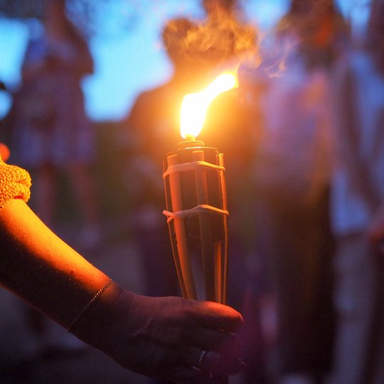 Stambiu planu matomas rankoje laikomas degantis deglas, kurio liepsna apšviečia sutemose vykstančią ekskursiją po Kalnų parką. Šilta ugnies šviesa ir dūmai sustiprina paslaptingą Vilniaus pilių legendų vakaro atmosferą.