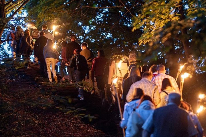 Naktinės ekskursijos dalyviai su deglais kyla mediniais laiptais Vilniaus pilių kalvomis, tarp medžių ir sutemų.