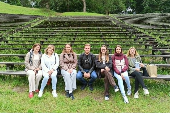 Grupė lankytojų sėdi ant lauko amfiteatro suolų žalioje Vilniaus pilių teritorijos aplinkoje. Už jų matyti medžiais apaugęs šlaitas ir žole apžėlę mediniai suolai.