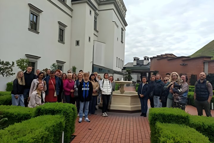 Ekskursijos dalyvių nuotrauka Vilniaus pilių teritorijoje, prie Valdovų rūmų. Matyti žmonių grupė kieme, žali gyvatvorių plotai, pastatų fasadai ir Gedimino kalno šlaitas fone.