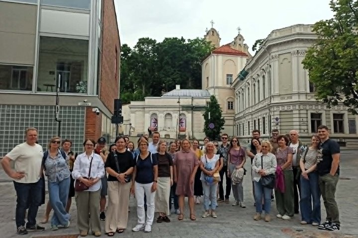 Ekskursijos dalyvių grupė stovi Vilniaus senamiestyje, šalia istorinių pastatų. Fone matyti bažnyčios bokštai, medžiai ir miesto architektūra.