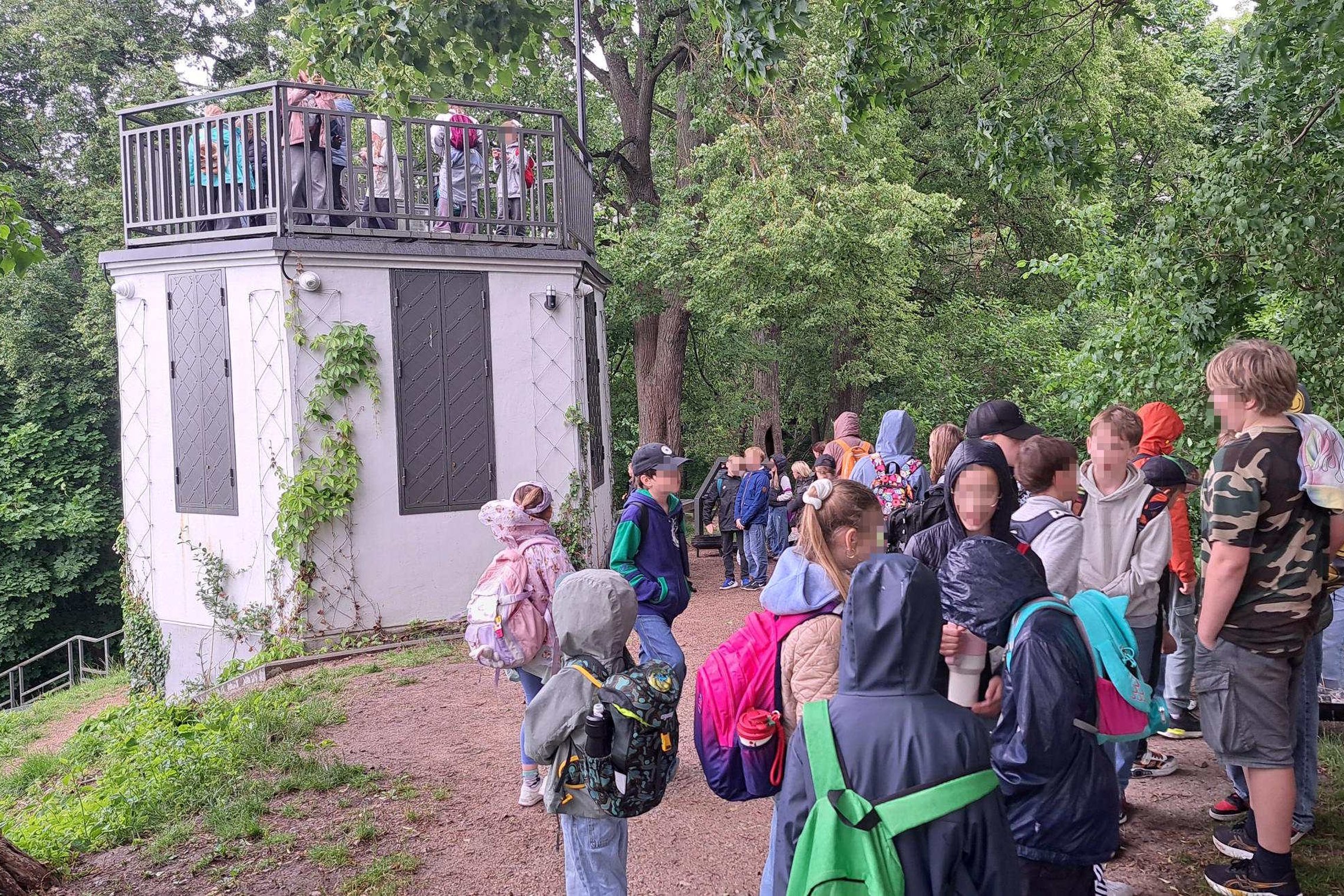 Grupė vaikų iš vasaros stovyklų dalyvauja edukacinėje veikloje Vilniaus pilių direkcijos teritorijoje. Vaikai, nešini kuprinėmis ir apsirengę lietui pritaikytais drabužiais, klausosi edukatoriaus pasakojimo medžių apsuptoje aikštelėje. Greta matoma apžval