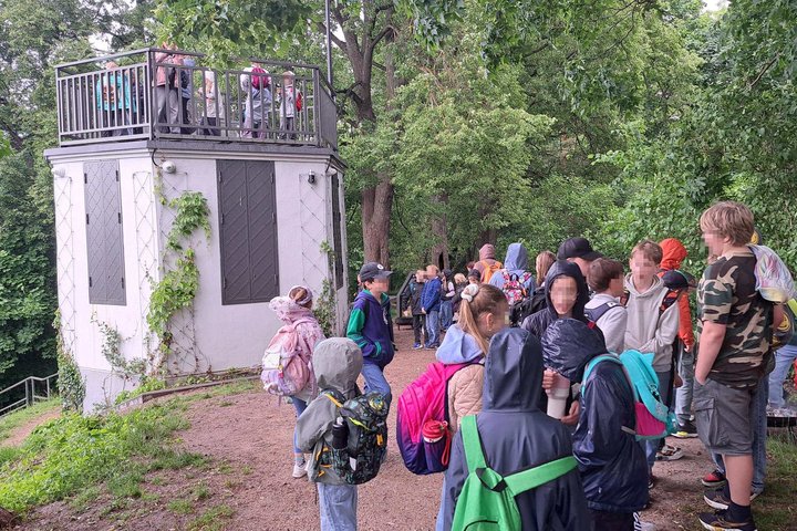 Grupė vaikų iš vasaros stovyklų dalyvauja edukacinėje veikloje Vilniaus pilių direkcijos teritorijoje. Vaikai, nešini kuprinėmis ir apsirengę lietui pritaikytais drabužiais, klausosi edukatoriaus pasakojimo medžių apsuptoje aikštelėje. Greta matoma apžval