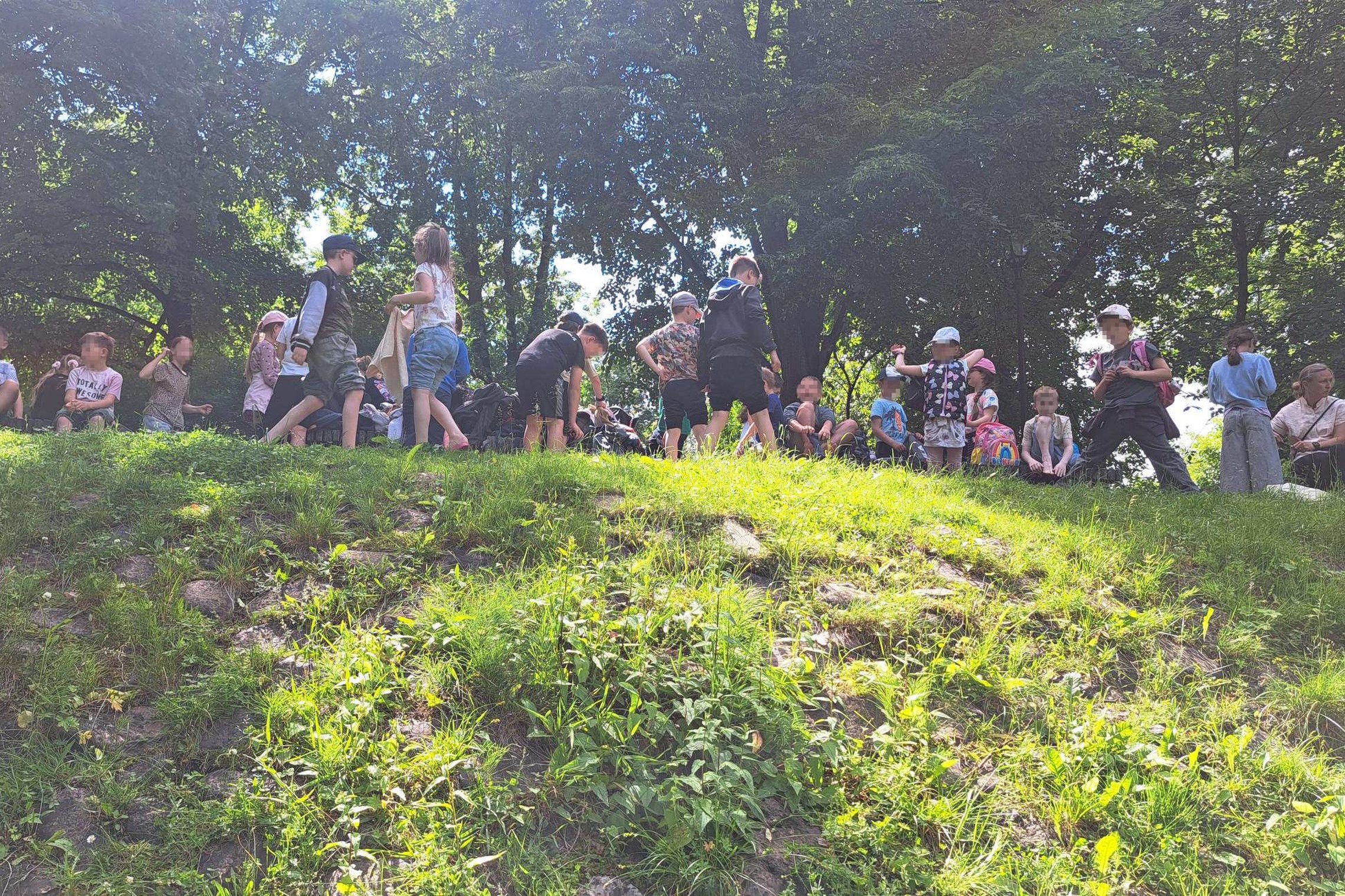Vaikų grupė dalyvauja edukacijoje gamtoje ant žolėto šlaito Vilniaus pilių teritorijoje. Mokiniai susibūrę medžių pavėsyje klausosi edukatoriaus, bendrauja tarpusavyje ir ruošiasi veikloms saulėtą vasaros dieną.