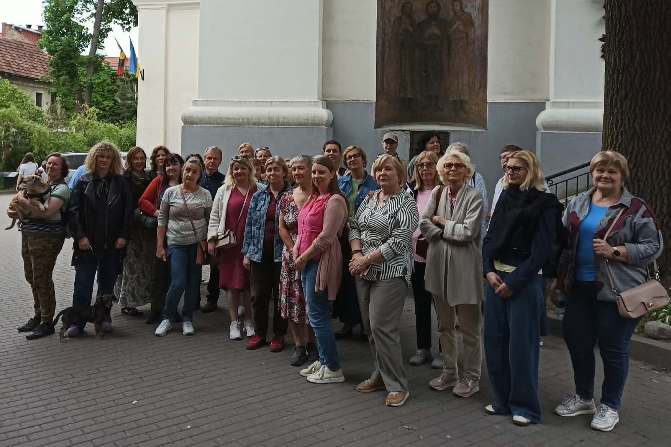 Grupė ekskursijos dalyvių stovi prie bažnyčios sienos Vilniaus senamiestyje. Virš įėjimo matyti religinis paveikslas, šalia plevėsuoja vėliavos.
