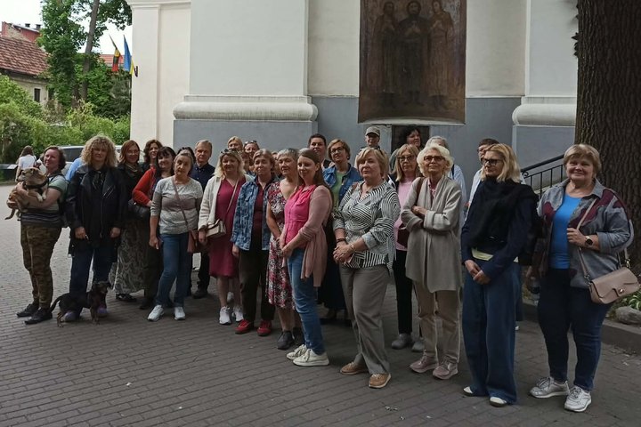 Grupė ekskursijos dalyvių stovi prie bažnyčios sienos Vilniaus senamiestyje. Virš įėjimo matyti religinis paveikslas, šalia plevėsuoja vėliavos.