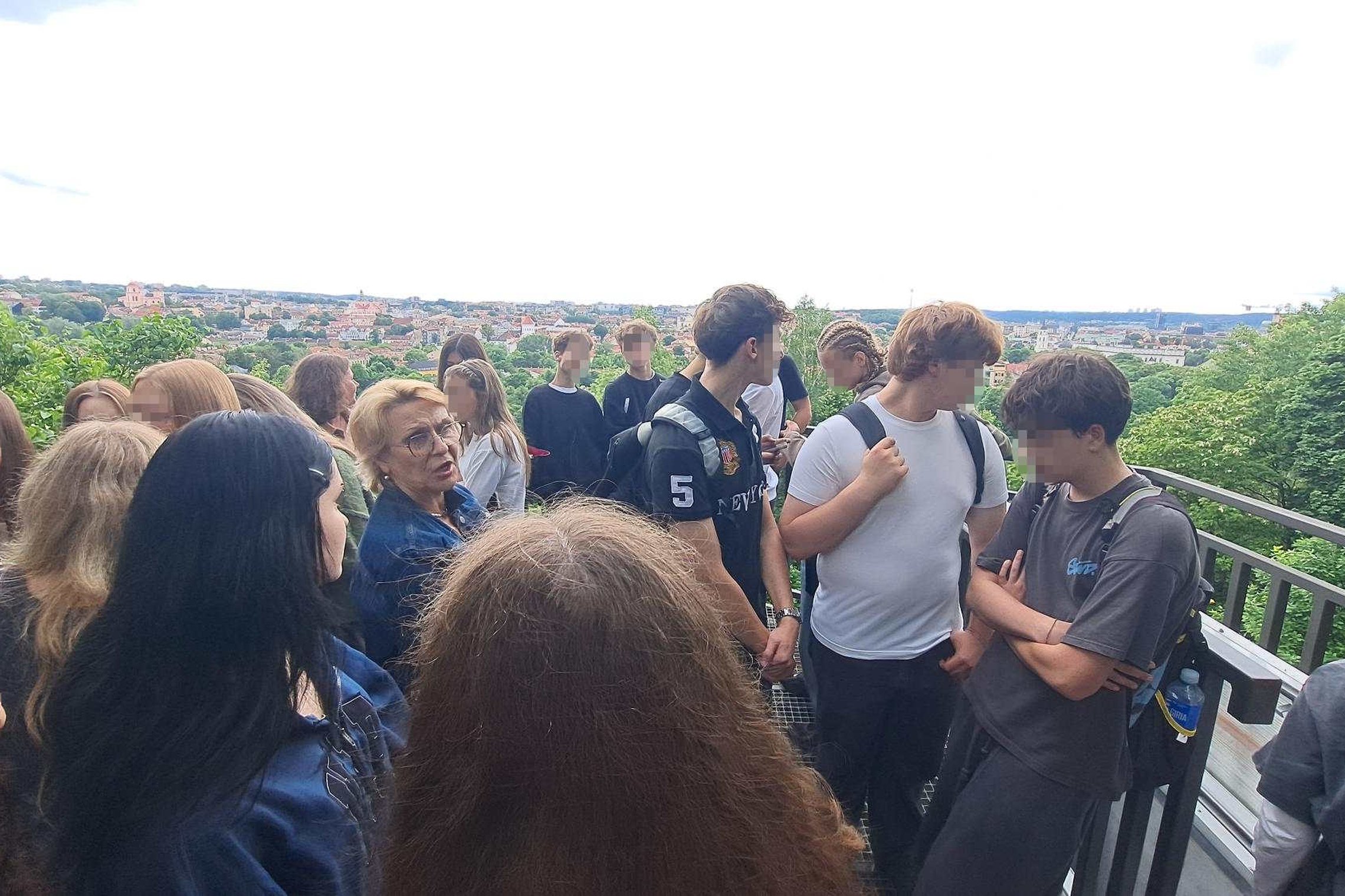 Vilniaus Užupio gimnazijos devintokai kartu su gide stovi apžvalgos aikštelėje Vilniaus pilių teritorijoje. Mokiniai klausosi pasakojimo apie miesto istoriją, o už jų atsiveria plati Vilniaus panorama su senamiesčio stogais ir žaliuojančiais upės slėnio š