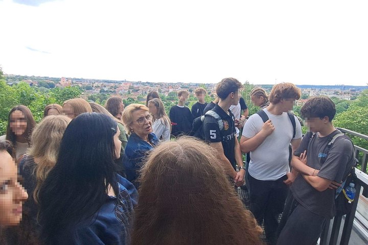 Vilniaus Užupio gimnazijos devintokai kartu su gide stovi apžvalgos aikštelėje Vilniaus pilių teritorijoje. Mokiniai klausosi pasakojimo apie miesto istoriją, o už jų atsiveria plati Vilniaus panorama su senamiesčio stogais ir žaliuojančiais upės slėnio š