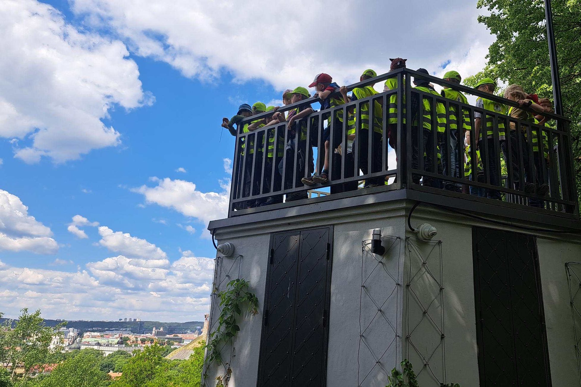 Nuotraukoje matyti vaikų grupė edukacinės veiklos metu apžvalgos aikštelėje, nuo kurios atsiveria Vilniaus panorama.