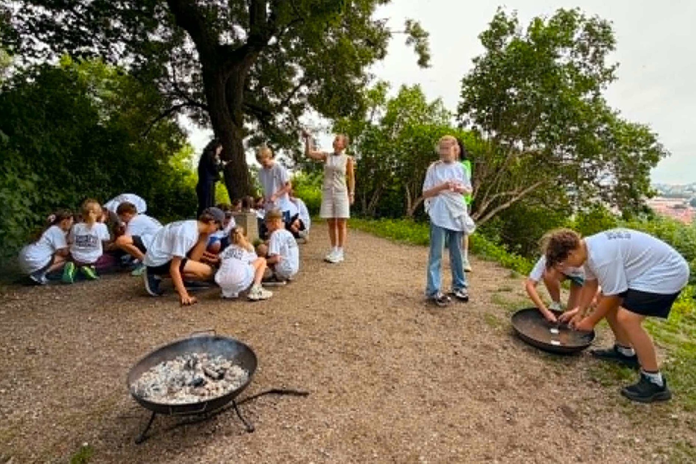 Vaikai dalyvauja edukacinėje veikloje Vilniaus pilių direkcijos teritorijoje – vieni tyrinėja užduotis prie laužavietės, kiti klausosi edukatorės pasakojimo po atviru dangumi. Fone matyti žaliuojantys medžiai ir Vilniaus panorama, o veiklos vyksta gamtos 