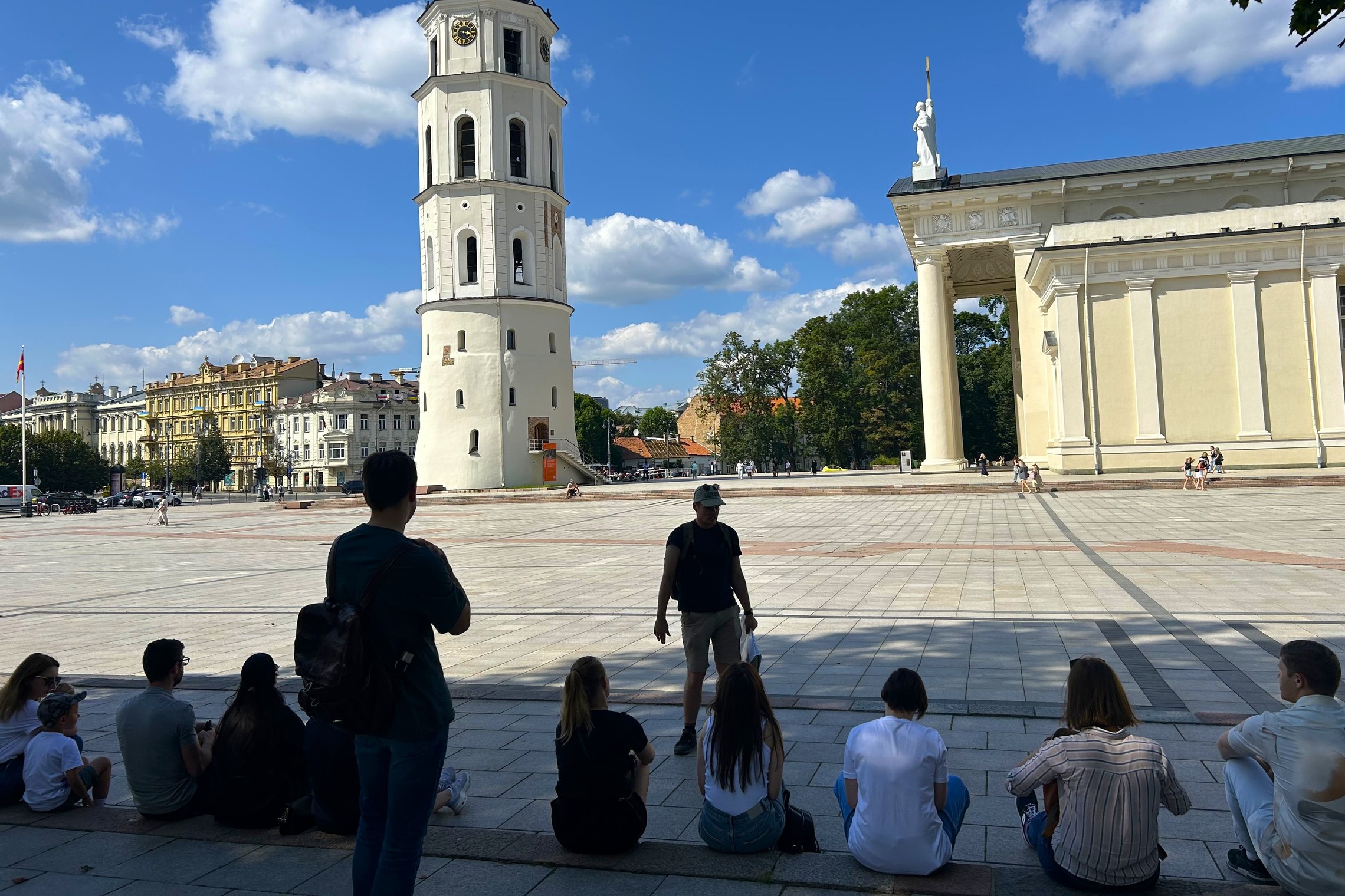 Ekskursijos dalyviai susėdę Katedros aikštėje klausosi gido pasakojimo, o fone matyti Vilniaus katedros varpinė ir Katedros pastatas. Saulėtą vasaros dieną grupė dalyvauja edukacinėje ekskursijoje Vilniaus pilių direkcijos teritorijoje.
