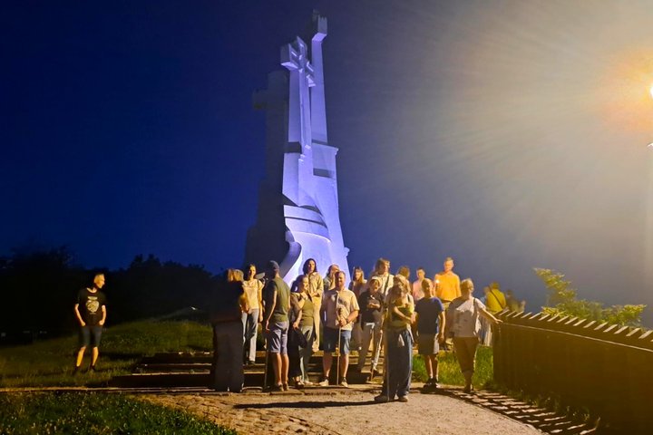Vakarinės ekskursijos dalyviai stovi prie Trijų Kryžių paminklo Vilniaus pilių direkcijos teritorijoje. Tamsų vasaros vakarą apšviestas paminklas ir susirinkusi žmonių grupė kuria paslaptingą atmosferą, primenančią Vilniaus pilių legendų pasakojimus.