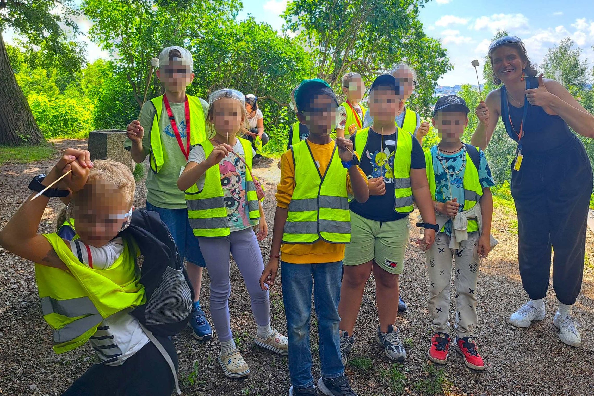 Grupė vaikų su ryškiomis saugos liemenėmis ir edukatorė pozuoja Vilniaus pilių direkcijos teritorijoje vykusios edukacijos metu. Vaikai rankose laiko edukacinėms veikloms skirtas priemones, o aplink matyti žaliuojanti gamta ir vasariška aplinka.