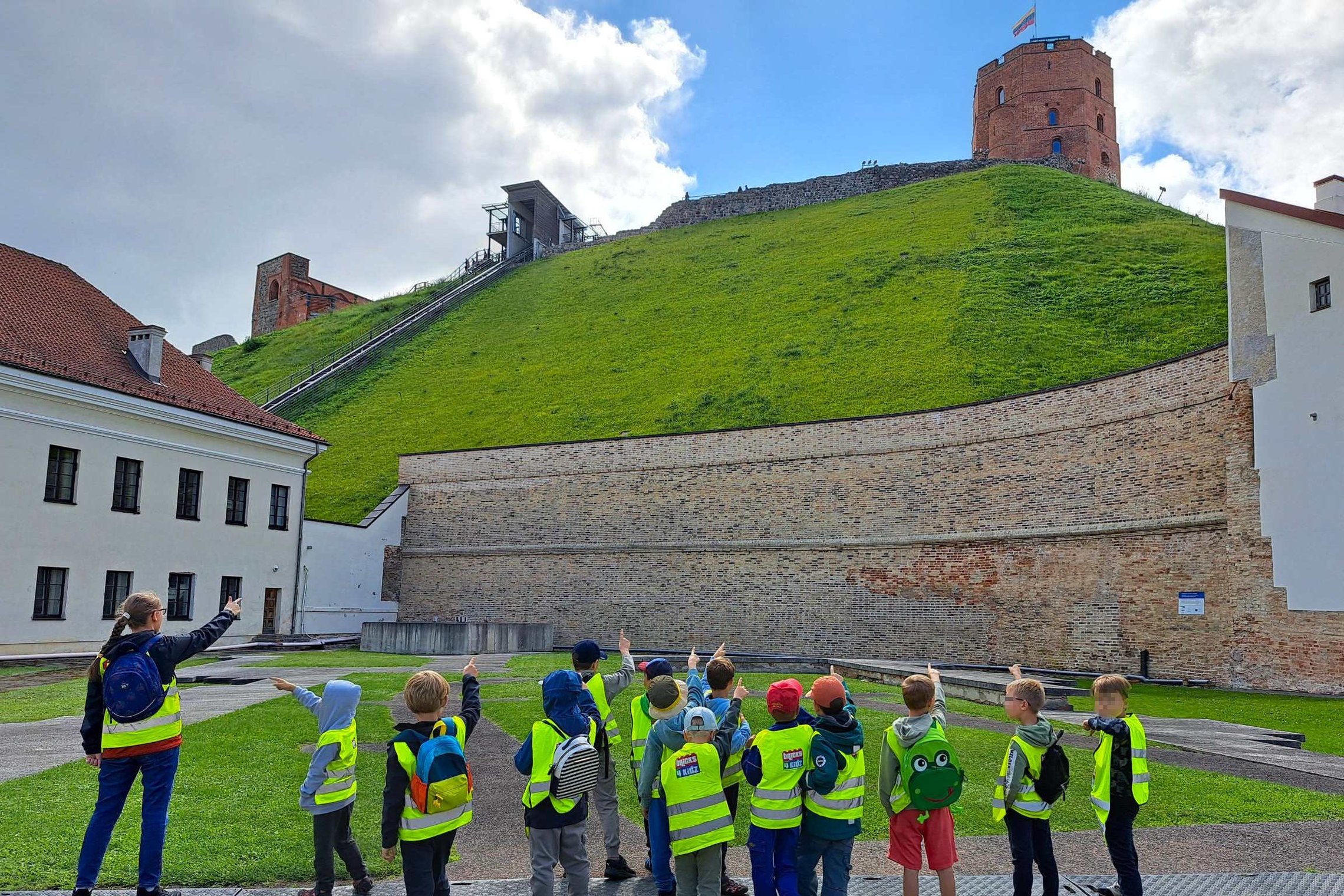 Vaikų grupė stovi Vilniaus pilių direkcijos teritorijoje ir rodo į Gedimino pilies bokštą ant Gedimino kalno. Visi vaikai, vilkintys ryškias apsaugines liemenes, žvelgia į kalną ir rankomis rodo bokšto kryptimi.