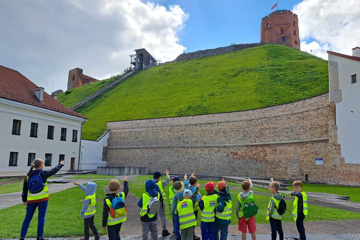 Vaikų grupė stovi Vilniaus pilių direkcijos teritorijoje ir rodo į Gedimino pilies bokštą ant Gedimino kalno. Visi vaikai, vilkintys ryškias apsaugines liemenes, žvelgia į kalną ir rankomis rodo bokšto kryptimi.