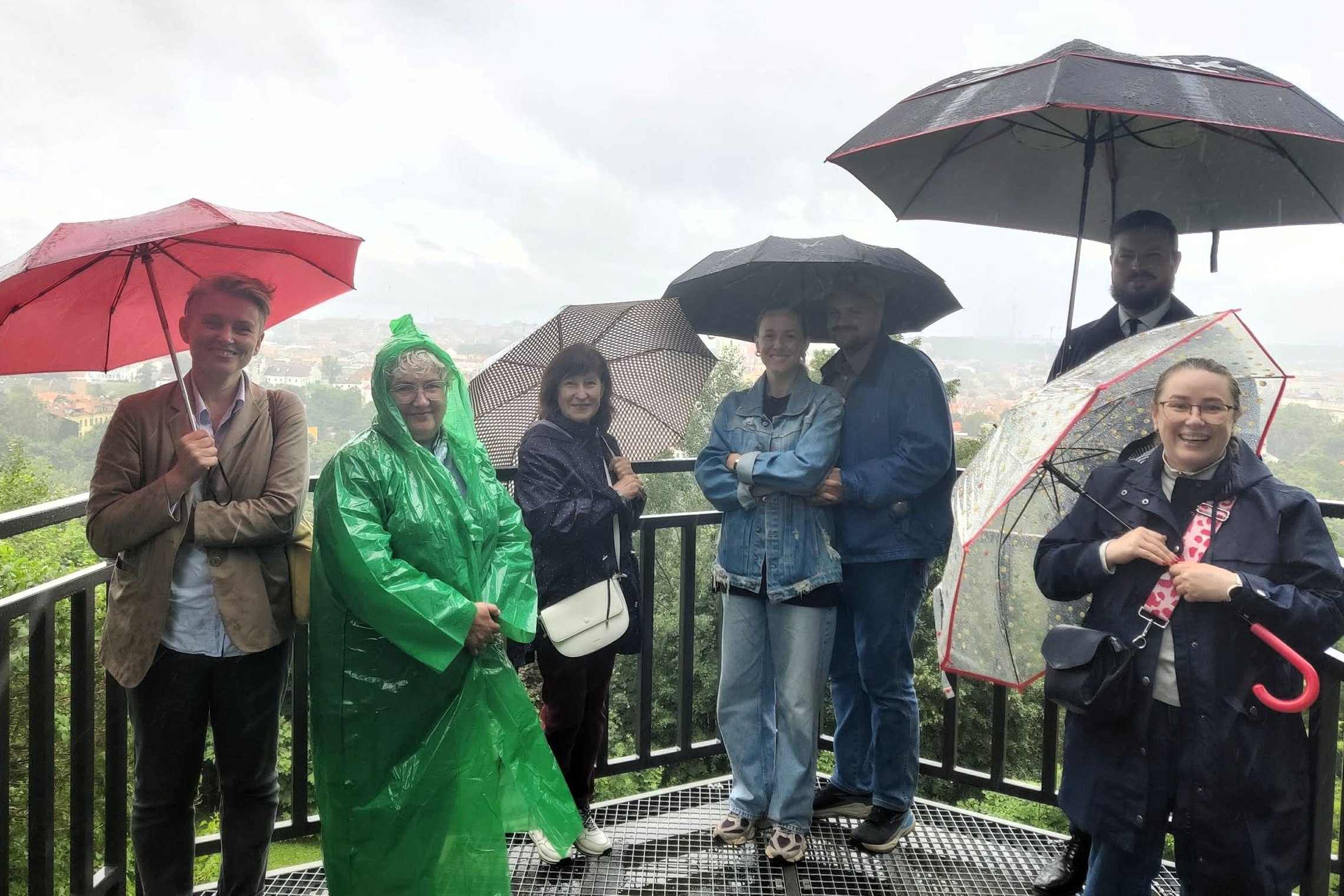 Ekskursijos dalyviai su skėčiais ir lietpalčiais ant Altanos kalno bokštelio lietingą šeštadienio rytą. Fone per rūką matyti Vilniaus panorama.