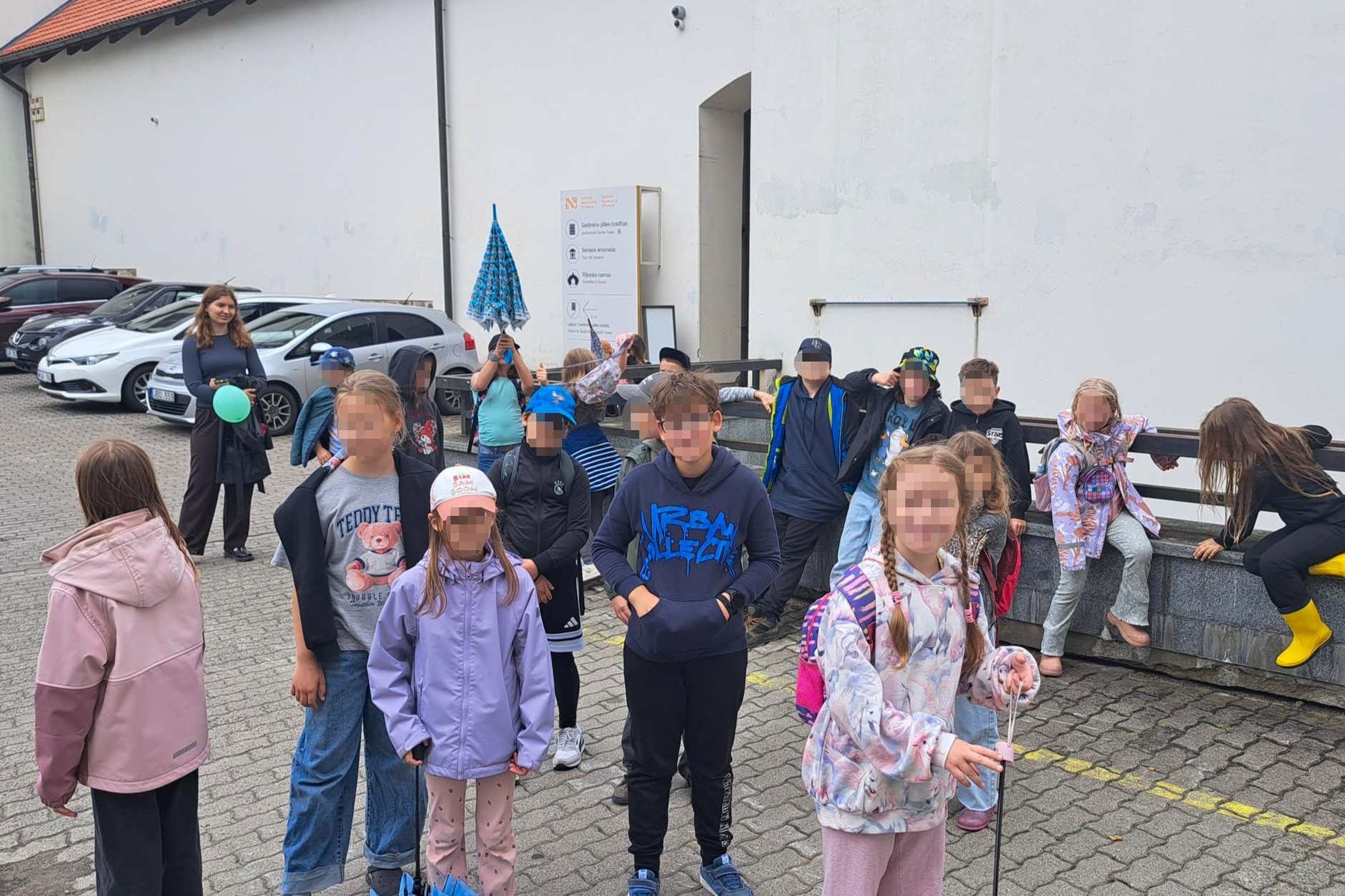 Vasaros stovyklos vaikai susirinkę Vilniaus pilių direkcijos kieme prieš edukaciją apie Vilniaus pilis, o grupę lydi edukatorė. Dalyviai stovi ir bendrauja lauko erdvėje prie istorinių pastatų, ruošdamiesi pažinčiai su Vilniaus pilių istorija.