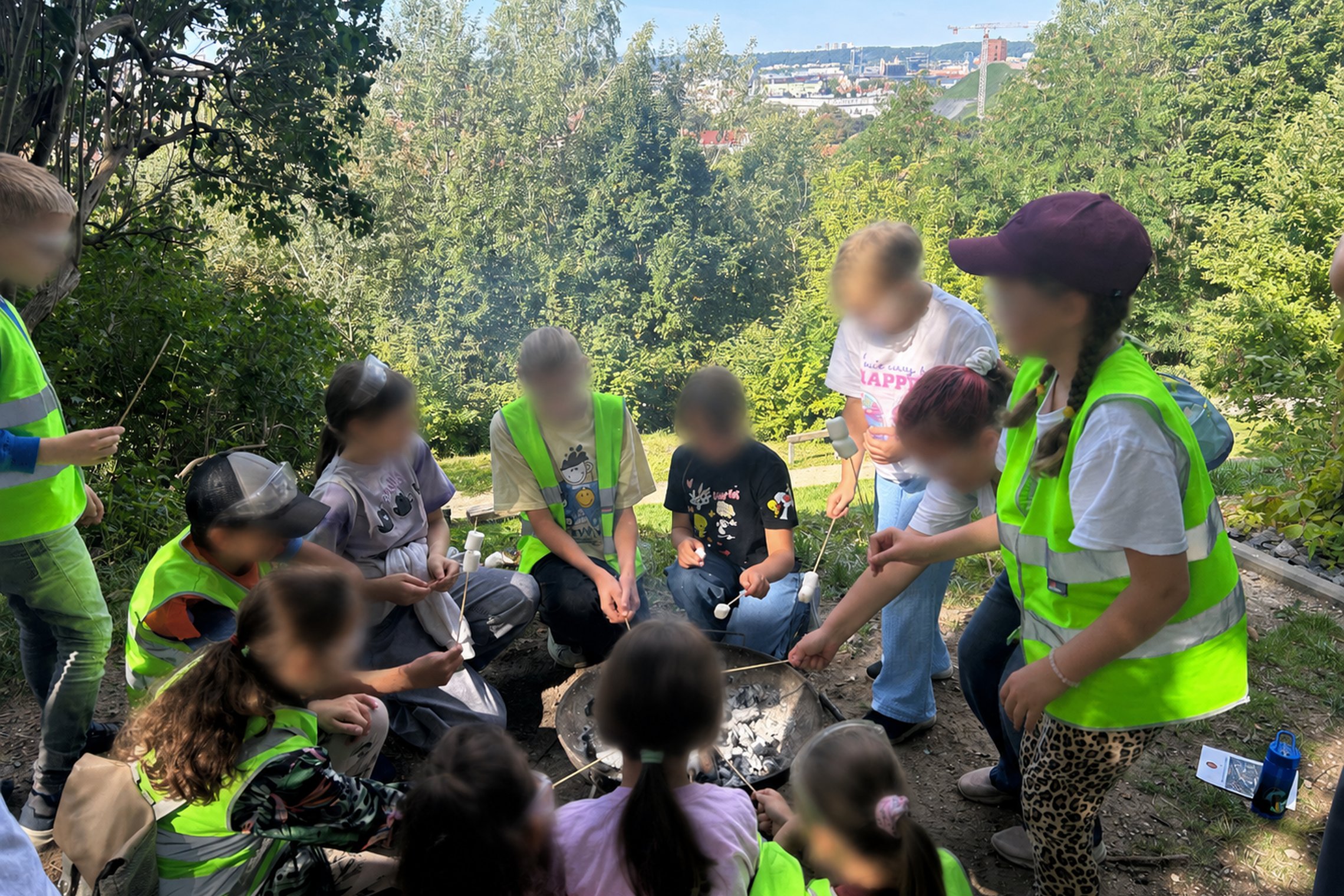 Vaikai su apsauginiais akiniais ir šviesą atspindinčiomis liemenėmis dalyvauja ugnies edukacijoje gamtos apsuptyje ant Altanos kalno – aplink laužavietę kepa zefyrus ant medinių iešmelių. Fone atsiveria žalia Vilniaus panorama.