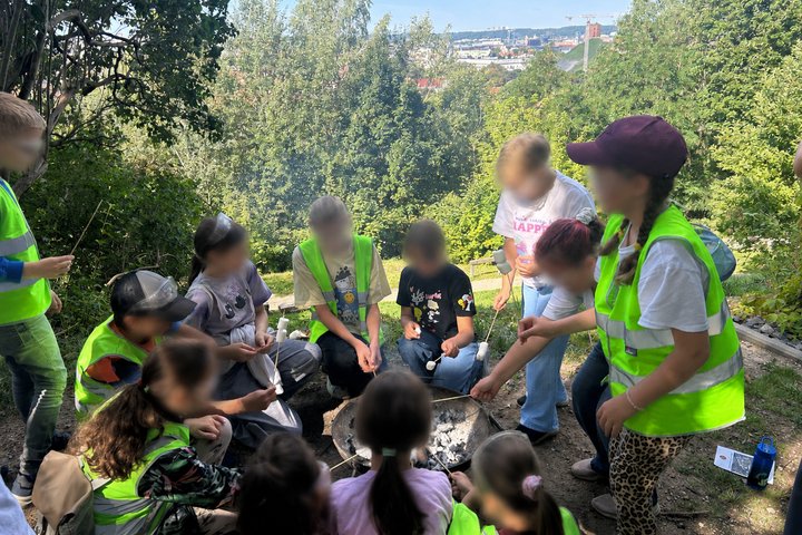 Vaikai su apsauginiais akiniais ir šviesą atspindinčiomis liemenėmis dalyvauja ugnies edukacijoje gamtos apsuptyje ant Altanos kalno – aplink laužavietę kepa zefyrus ant medinių iešmelių. Fone atsiveria žalia Vilniaus panorama.