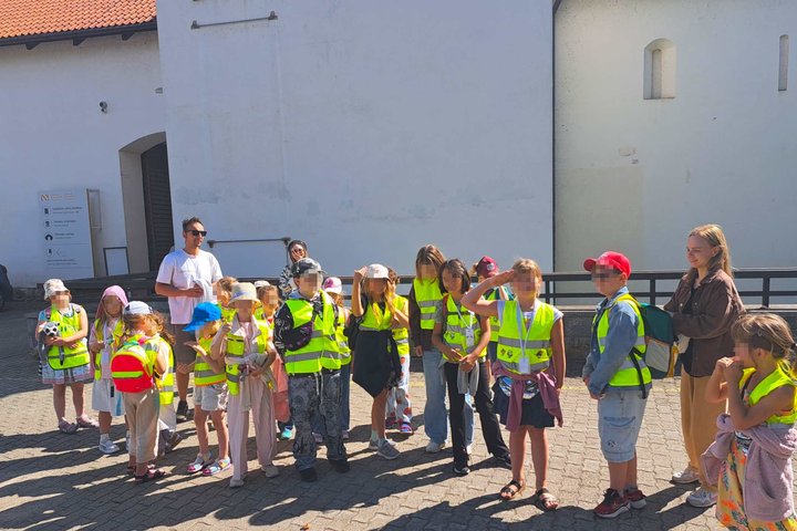 Grupė vaikų su ryškiaspalvėmis saugos liemenėmis stovi Vilniaus pilių teritorijoje prie istorinio pastato. Kartu su vaikais matomi edukacijos vadovai, dalyviai klausosi pasakojimo ir ruošiasi tęsti pažintį su Vilniaus pilių istorija.
