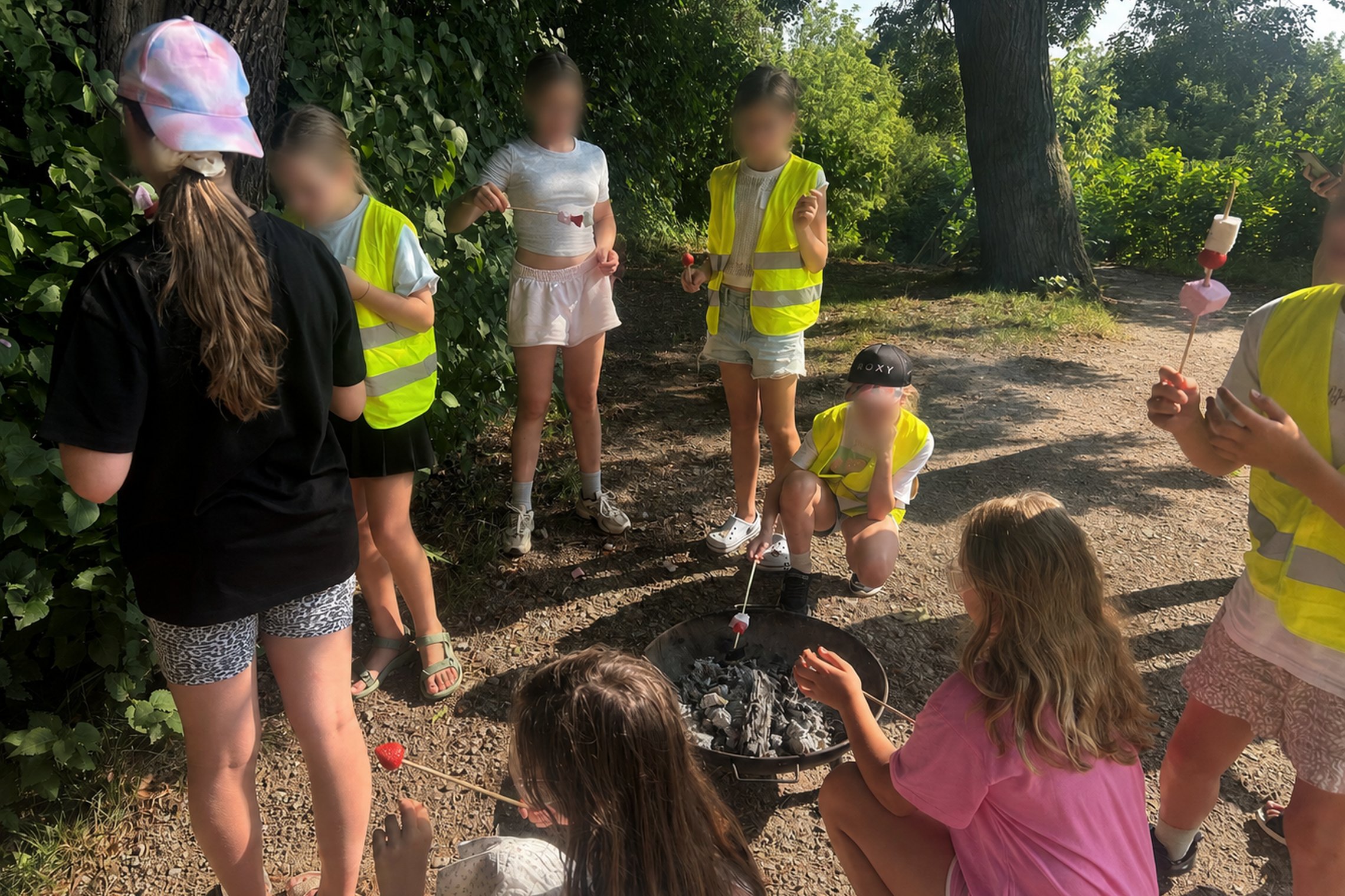 Vaikai, apsirengę geltonomis liemenėmis, ant Altanos kalvos dalyvauja ugnies edukacijoje – susibūrę aplink nedidelį laužą jie ant medinių iešmelių kepa zefyrus.