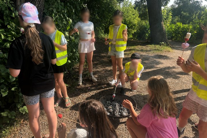 Vaikai, apsirengę geltonomis liemenėmis, ant Altanos kalvos dalyvauja ugnies edukacijoje – susibūrę aplink nedidelį laužą jie ant medinių iešmelių kepa zefyrus.