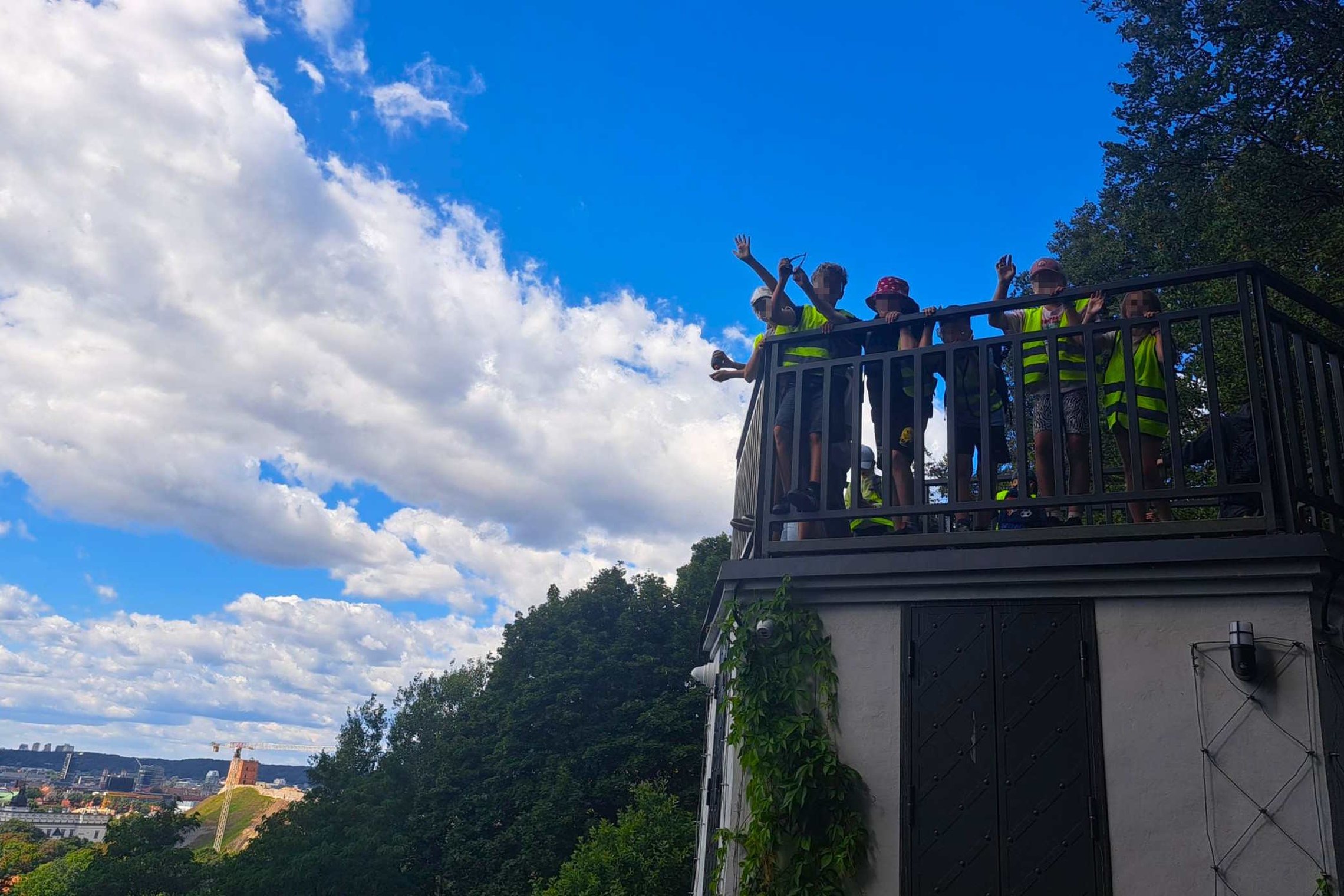 Altanos bokštelio apžvalgos aikštelėje stovi grupė vasaros stovyklos dalyvių su ryškiaspalvėmis liemenėmis, dalis jų mojuoja. Fone matyti Vilniaus miesto panorama, žaliuojantys medžiai ir mėlynas dangus su baltais debesimis.