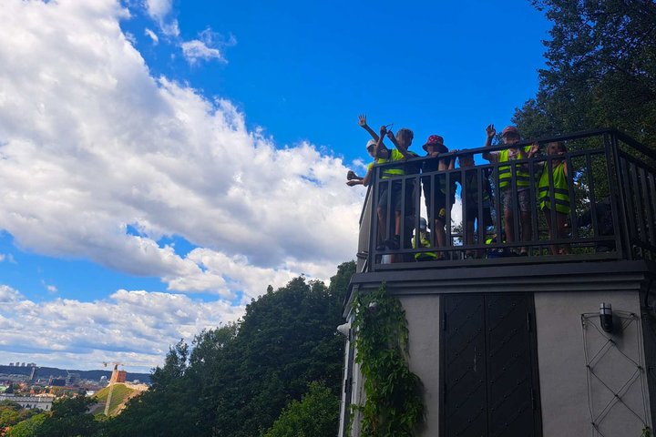 Altanos bokštelio apžvalgos aikštelėje stovi grupė vasaros stovyklos dalyvių su ryškiaspalvėmis liemenėmis, dalis jų mojuoja. Fone matyti Vilniaus miesto panorama, žaliuojantys medžiai ir mėlynas dangus su baltais debesimis.