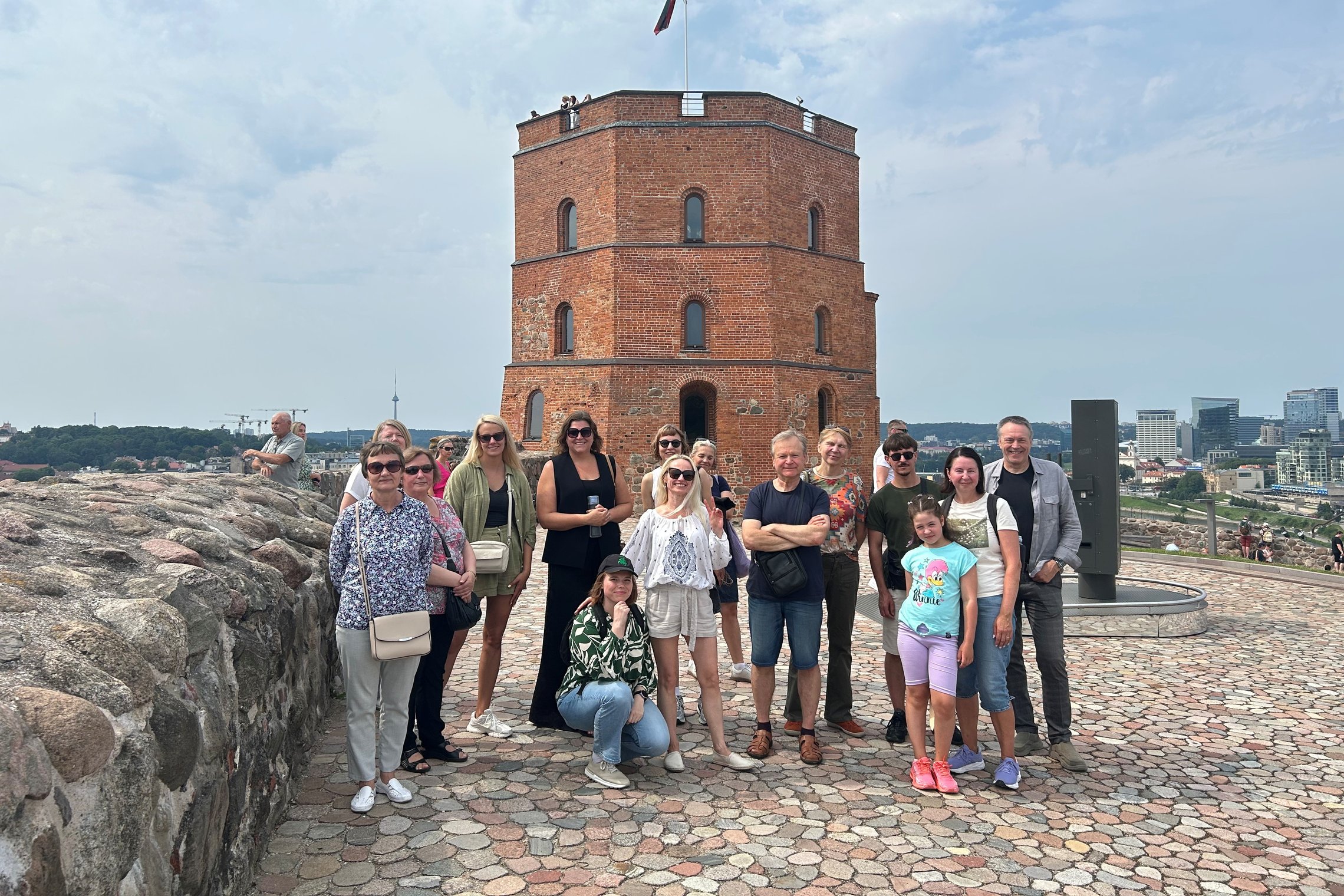 Grupė ekskursijos „Vilniaus pilių legendos“ dalyvių pozuoja prie Gedimino pilies bokšto ant Gedimino kalno. Už jų matyti Gedimino pilies bokštas, Lietuvos vėliava ir Vilniaus miesto panorama.