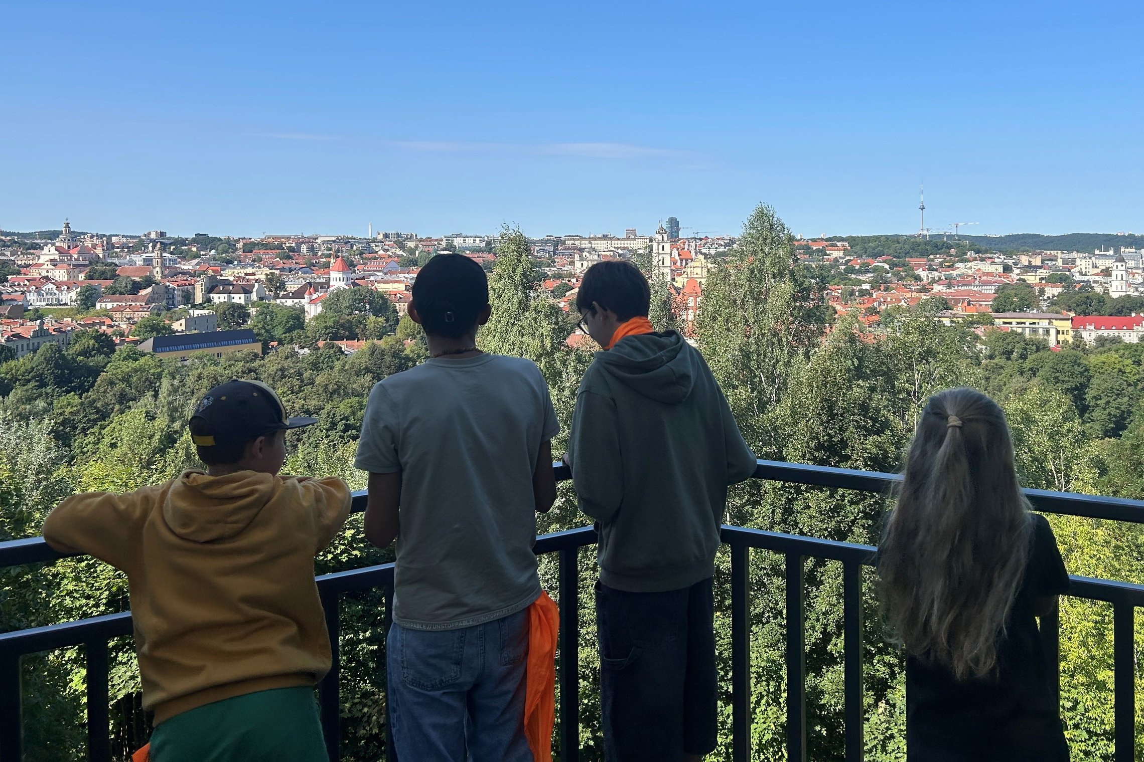 Keturi vaikai, nufotografuoti iš nugaros, stovi prie metalinės aptvaro tvorelės Altanos bokštelyje žiūri į Vilniaus senamiesčio panoramą po giedru, melsvu dangumi.