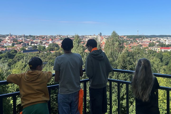 Keturi vaikai, nufotografuoti iš nugaros, stovi prie metalinės aptvaro tvorelės Altanos bokštelyje žiūri į Vilniaus senamiesčio panoramą po giedru, melsvu dangumi.