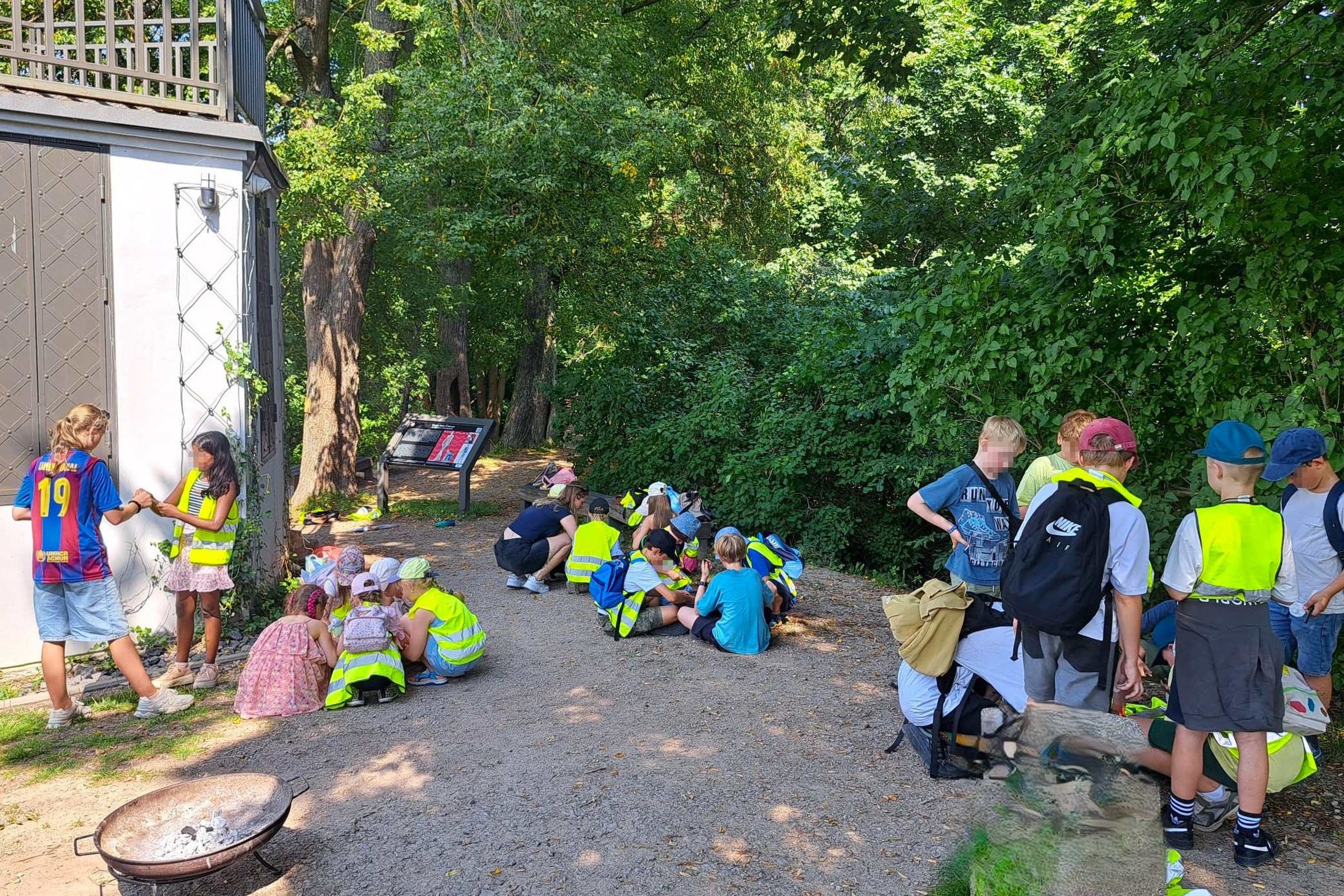 Vaikai, vilkintys ryškiaspalves liemenes, dalyvauja edukacinėje veikloje žalumos apsuptoje Vilniaus pilių teritorijoje. Dalyviai susibūrę į grupeles atlieka užduotis, o pirmame plane matoma ugniavietė.