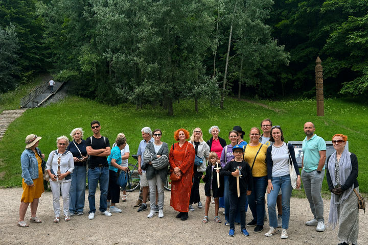 Grupė ekskursijos „Vilniaus pilių legendos“ dalyvių fotografuojasi prie Bekešo kalno Vilniuje. Už jų atsiveria žaliuojanti kalvos aplinka, medinis takas ir istorinis ženklas, o šalia matomi informaciniai stendai.